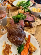 A close-up, slightly high-angle shot of a generous Sunday roast sharing platter featuring multiple meats, Yorkshire puddings, and various vegetable sides, accompanied by a glass of rosé wine.