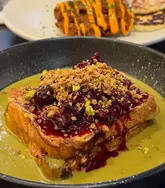 A close-up shot of a plate of French toast topped with berries and pistachios, served with a green sauce. In the background, a plate of fried chicken and halloumi is partially visible.