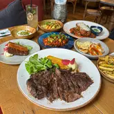 A high-angle, flat-lay photograph of a table spread at Sonrisa restaurant, featuring a large, well-seared steak in the foreground, accompanied by various small plates, sides, and a cocktail.