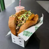 A high-quality, close-up shot of a loaded breakfast-style sandwich served in branded takeaway packaging, accompanied by a pink drink in the background. The lighting is soft and natural, highlighting the textures of the bread, egg, and sauce.