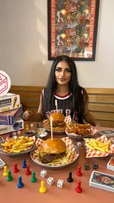 A promotional lifestyle shot featuring a person at a table in a board game cafe, surrounded by burgers, fries, and various game components, effectively showcasing the venue's unique selling point.