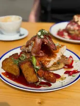 A close-up, eye-level shot of a brunch dish featuring fried chicken, bacon, and syrup, served on a white plate with a blue rim. A coffee cup and another plate are visible in the blurred background.