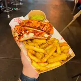 A first-person, handheld shot of a street food meal consisting of two chicken tacos, a side of fries, and a small portion of guacamole, served in a cardboard tray. The image is taken in a dimly lit environment, likely a food hall, using a direct camera flash.