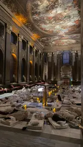 A cinematic montage showcasing a 'Lie Down and Listen' wellness event held in the historic Painted Hall in London. The video features slow, sweeping shots of the grand architecture, attendees lying on mats, and performers playing violin and singing bowls, creating a tranquil and immersive atmosphere.