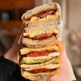 A high-quality, close-up shot of three stacked sandwiches being held by a person. The image effectively showcases the fresh ingredients, textures, and layers of the food, making it highly appealing for a lunch or brunch setting. The shallow depth of field keeps the focus entirely on the sandwiches.
