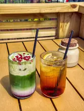 A bright, sunny shot of two tall, ribbed glasses containing a matcha latte and an iced tea, placed on a wooden outdoor table. The image captures a casual, refreshing moment at a venue that doubles as a florist.