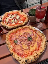 A top-down shot of two pizzas and a cocktail on an outdoor wooden table, capturing a casual dining experience.