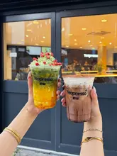 A bright, clear promotional shot featuring two hands holding colourful drinks in front of a Teapresso storefront, effectively showcasing the product and the venue's branding.