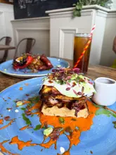 A close-up, eye-level shot of a chicken and waffle dish served on a blue plate, featuring a stack of waffles topped with crispy fried chicken, a fried egg, and microgreens, all drizzled with a vibrant orange sauce. A drink and another plate are visible in the background.