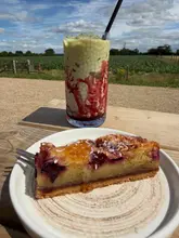 A close-up shot of an iced raspberry matcha drink and a slice of cherry bakewell cake served on a wooden table at a farm shop cafe, with a rural field in the background.