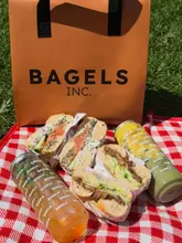 A bright, overhead shot of a picnic spread featuring two bagels and two drinks from Bagels Inc, arranged on a red and white checkered blanket on grass.