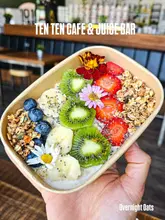 A vibrant, high-quality close-up shot of a bowl of overnight oats topped with fresh kiwi, strawberries, blueberries, banana, and edible flowers. The image is held by a hand, providing a sense of scale and human connection, with bright, natural lighting that highlights the freshness of the ingredients.