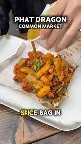 A close-up, top-down action shot of a spice bag being drizzled with curry sauce, highlighting the crispy chicken, chips, and peppers in a takeaway tray.