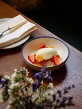 A high-quality, professional food photograph featuring a plated dessert, likely a fruit-based dish with a scoop of ice cream or sorbet, served in a ceramic bowl on a wooden table. The composition uses a shallow depth of field, with out-of-focus flowers in the foreground adding a soft, natural frame to the subject.