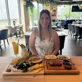 A promotional image featuring a person sitting at a restaurant table with a spread of food including a salmon fillet, steak, various sides, and two cocktails.