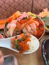 A close-up, first-person perspective shot of a spoonful of Japanese donburi, featuring fresh fish and ikura, held in front of the main bowl.