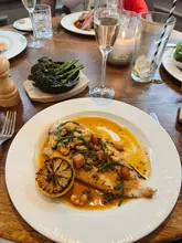A top-down shot of a restaurant table featuring a main dish of Dover sole with mussels, capers, and samphire, accompanied by a side of broccolini and a glass of sparkling wine.