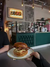 A casual, first-person perspective shot of a bacon and egg brioche bun held in front of a cafe counter. The image captures the atmosphere of the venue, with the menu board and coffee equipment visible in the background.