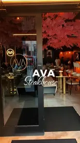 A clean, eye-level shot of the entrance to Ava Steakhouse, showcasing the modern, moody interior with warm lighting and decorative cherry blossom trees.
