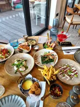 A high-quality, overhead flat-lay shot of a restaurant table spread featuring a variety of small plates, skewers, and cocktails. The lighting is bright and natural, evoking a summer dining atmosphere, with the table set against a backdrop of a restaurant patio or deck.
