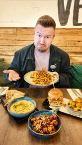 A creator visits Vegivores in Caversham to review their plant-based menu. He samples several dishes, including hummus, 'drunken knots', a mac and cheese waffle, and a burger, providing commentary on the flavors and textures while discussing the restaurant's unique offering in the area.