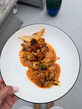 A high-quality, overhead shot of a plate of pan-fried dumplings drizzled with chili oil, held by a person outdoors. The lighting is natural and even, highlighting the texture of the dumplings and the vibrant color of the sauce.