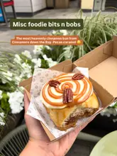 A close-up, handheld shot of a pecan caramel cinnamon bun in a takeaway box, set against a soft-focus outdoor background.