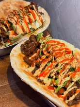 A close-up, top-down angled shot of two spicy beef rotis served in metal trays on a wooden surface. The focus is sharp on the front roti, highlighting the texture of the beef and the drizzle of sauces, though the lighting is noticeably harsh.