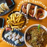 A flat-lay overhead shot of a spread of Japanese-inspired dishes including gyoza, bao buns, fries, sushi, and stir-fried noodles, served on a wooden table.