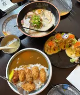 A high-quality, overhead flat-lay shot of a Japanese meal spread, featuring ramen, curry with fried oysters, and sushi rolls. The lighting is soft and natural, highlighting the textures and colors of the food effectively.