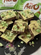 A close-up, overhead shot of a homemade Aero Mint traybake, featuring squares of chocolate biscuit base topped with white chocolate and Aero Mint bubbles, served on a slate board.