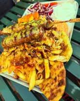 A high-angle, close-up shot of a large Greek food platter featuring chicken gyros, pork souvlaki, fried halloumi, fries, and pitta bread, served on a metal tray on a green slatted table.