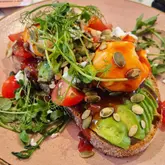 A close-up, high-angle shot of avocado and feta toast topped with poached eggs, sweet chilli sauce, and pumpkin seeds, served at a play cafe.