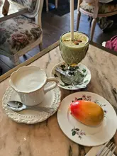 A top-down shot of a table at a cafe featuring a mango-shaped mousse cake, a green avocado-based drink, and a cup of coffee, served on floral china.