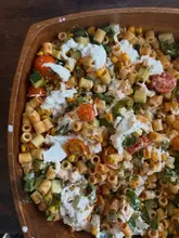A top-down, close-up shot of a homemade pasta salad served in a wooden bowl, featuring pasta, cherry tomatoes, cucumber, corn, and dollops of burrata cheese.