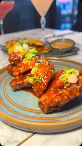 A montage of food and drink items served at a restaurant, including cocktails, Korean wings, chicken satay, lamb chops, fried rice, and cheesecake, followed by shots of the venue's interior and outdoor seating area.