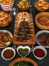 A professional, overhead flat-lay photograph of a large BBQ catering spread, featuring skewers, burgers, corn, and various salads, arranged neatly on a dark outdoor table.