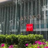 A clean, professional eye-level shot of the exterior of Akoko restaurant, featuring the brand signage and a Michelin 2026 plaque, framed by greenery in the foreground.