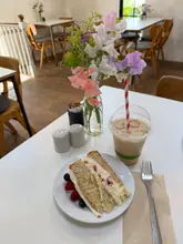 A bright, natural-light shot of a slice of berry cake and an iced latte at a cafe, featuring fresh flowers on the table.