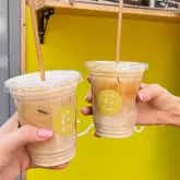 A bright, cheerful shot of two iced coffees being held up against a vibrant yellow wall, likely taken outside a cafe. The image focuses on the product, using the brand's color scheme to create a cohesive and appealing visual.