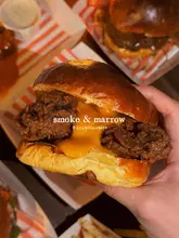 A close-up, handheld shot of a fried chicken burger with melted cheese, captured in a casual, dimly lit venue setting. The image uses direct flash to highlight the texture of the crispy chicken and the gooey cheese, effectively showcasing the food's appeal for social media.