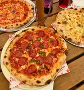 A top-down shot of three pizzas served on metal trays on a wooden outdoor table, accompanied by drinks. The image captures a casual dining experience, likely at an outdoor venue.