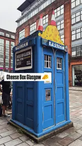 A short, punchy reel featuring a street food stall operating out of a converted police box in Glasgow. The video focuses on the preparation of raclette cheese toasties, showing the cheese being melted and scraped, and includes shots of the unique outdoor seating in a clear dome tent.