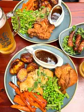 A high-quality, overhead flat-lay photograph of two Sunday roast dinners served at a pub. The image features plates filled with roast meat, vegetables, potatoes, and Yorkshire puddings, accompanied by individual gravy boats and a side of pigs in blankets. A glass of beer is visible on the side, contributing to the casual, social atmosphere of the venue.