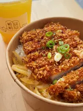 A close-up, high-angle shot of a pork katsu don bowl served in a takeaway container, accompanied by a drink in the background. The image highlights the crispy texture of the breaded pork cutlet and the appetizing presentation of the dish.