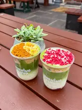 A bright, well-composed close-up shot of two layered matcha drinks in clear plastic cups, set on a wooden outdoor table. The drinks are topped with distinct garnishes, and the background features a soft-focus outdoor seating area with a small plant, creating a clean and inviting aesthetic.
