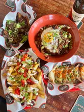 A high-angle, flat-lay shot of a spread of Asian-inspired street food dishes including wings, loaded fries, dumplings, and a noodle bowl, accompanied by a bottle of beer on a wooden table.