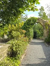A scenic, eye-level shot of a gravel path leading through a stone archway covered in ivy at a garden centre, surrounded by lush greenery and trees under a bright, sunny sky.