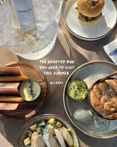 A flat-lay shot of a table at a rooftop bar featuring a burger, salad, focaccia, churros, and a bottle of wine. The image uses a top-down perspective to showcase the spread.