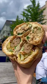 A close-up, first-person shot of two halves of a hearty, potato-stuffed sandwich being held outdoors.